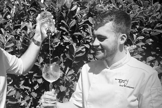 Two chefs pouring a glass of wine in front of bushes.
