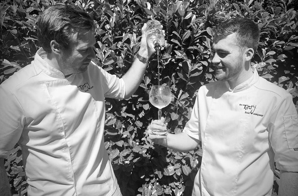 Two chefs holding wine glasses in front of bushes.